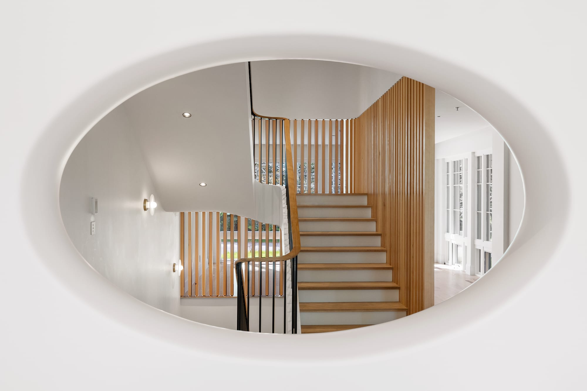 The stair hall through its oval opening, oak slat wall against painted risers.