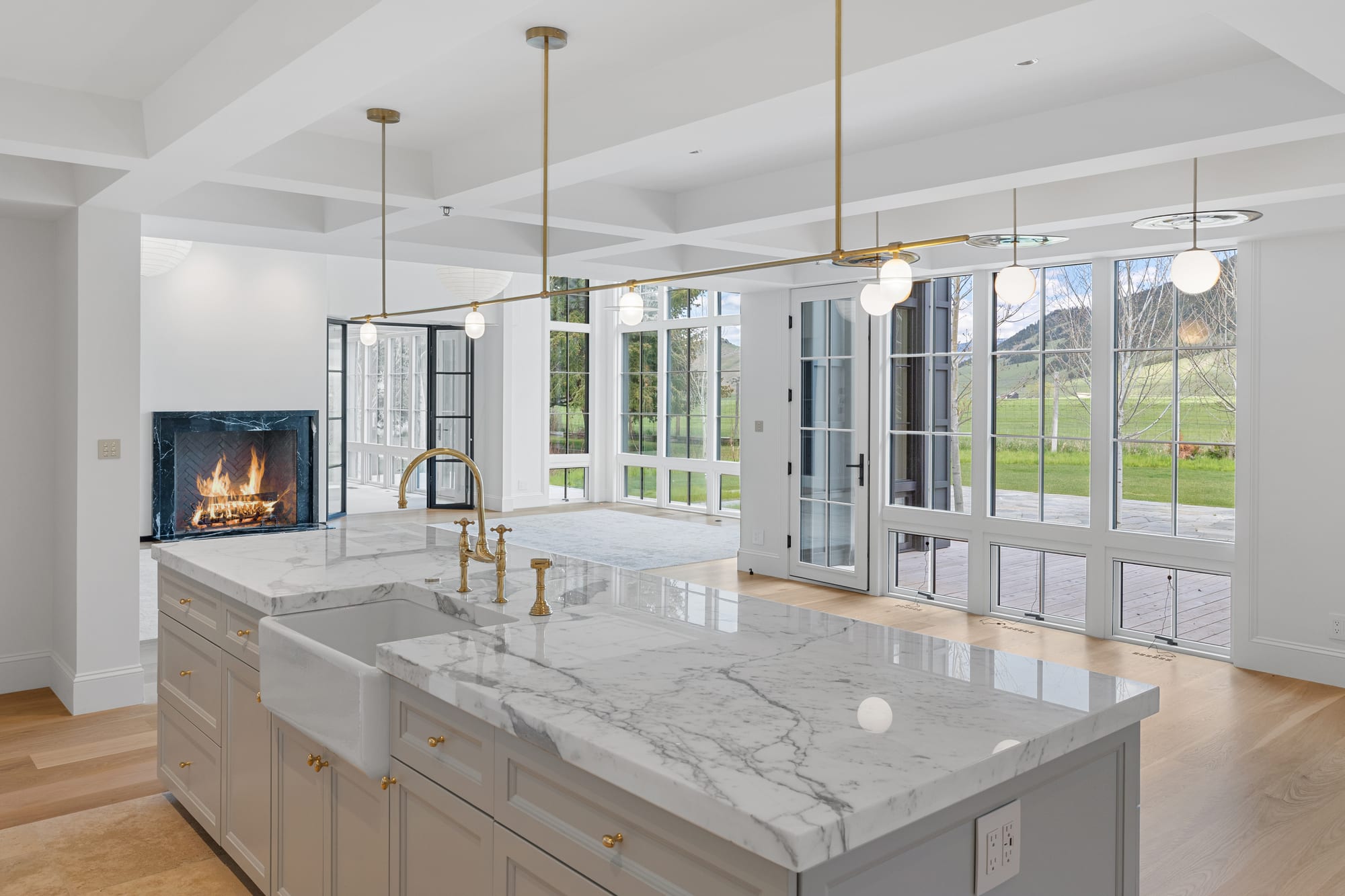 Kitchen island in marble with farmhouse sink, brass chandelier and hearth beyond.